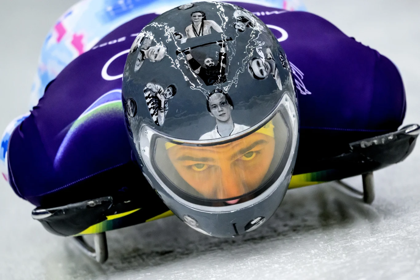 Władyslaw Hersakiewicz Zawodnik skeletonowy w trakcie jazdy po lodowym torze w sportowym stroju, jego kask ozdobiony grafikami przedstawiającymi twarze różnych osób i motywy artystyczne, skupiony wzrok sportowca widoczny przez wizjer.