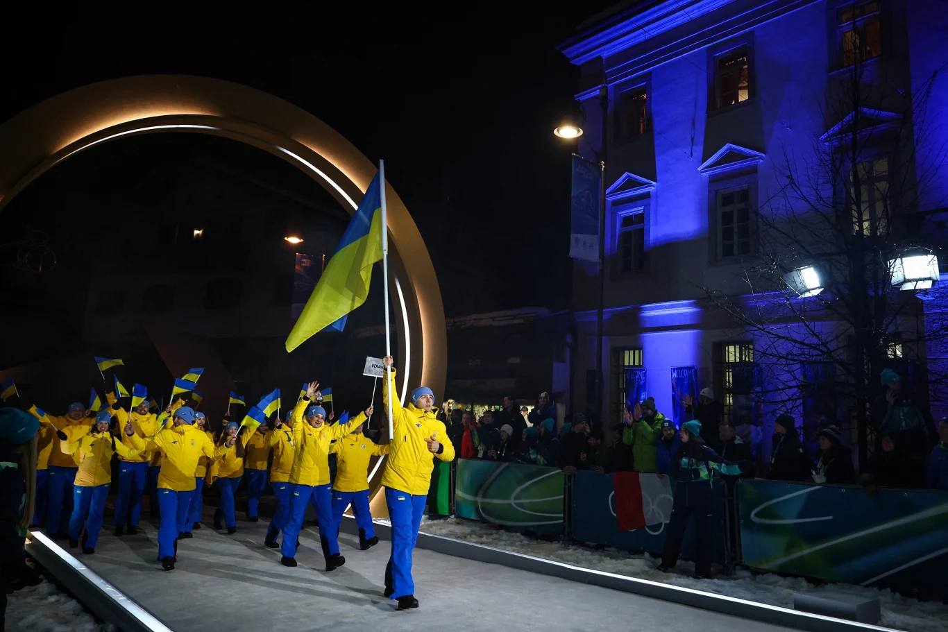 Władyslaw Hersakiewicz z ukraińską flagą Grupa sportowców ubranych w żółto-niebieskie stroje maszeruje z flagą Ukrainy podczas oficjalnej ceremonii, otoczona przez widzów i efektowne oświetlenie budynku w tle.