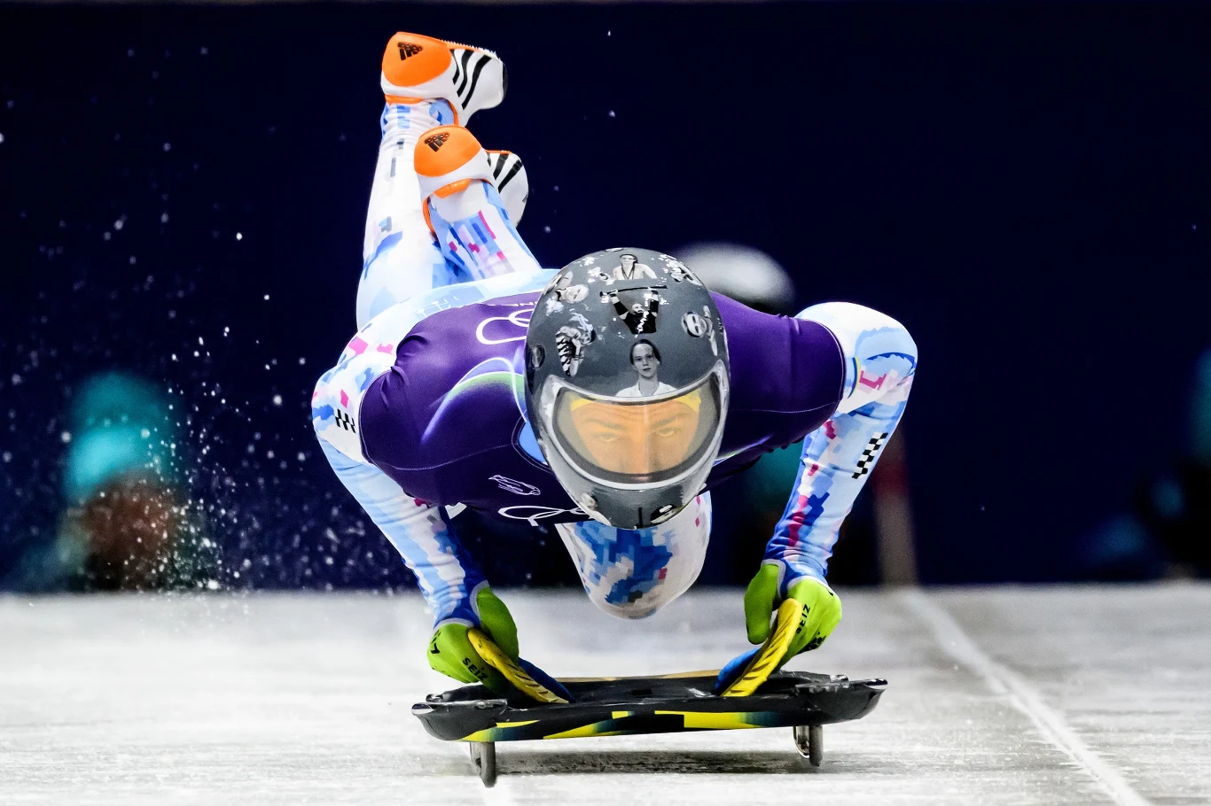 Władyslaw Hersakiewicz Zawodnik w kolorowym stroju sportowym ślizga się na skeletonie po zlodowionym torze, dynamiczna poza z uniesionymi nogami i skupioną twarzą, wyraźne emocje oraz energia rywalizacji sportowej.