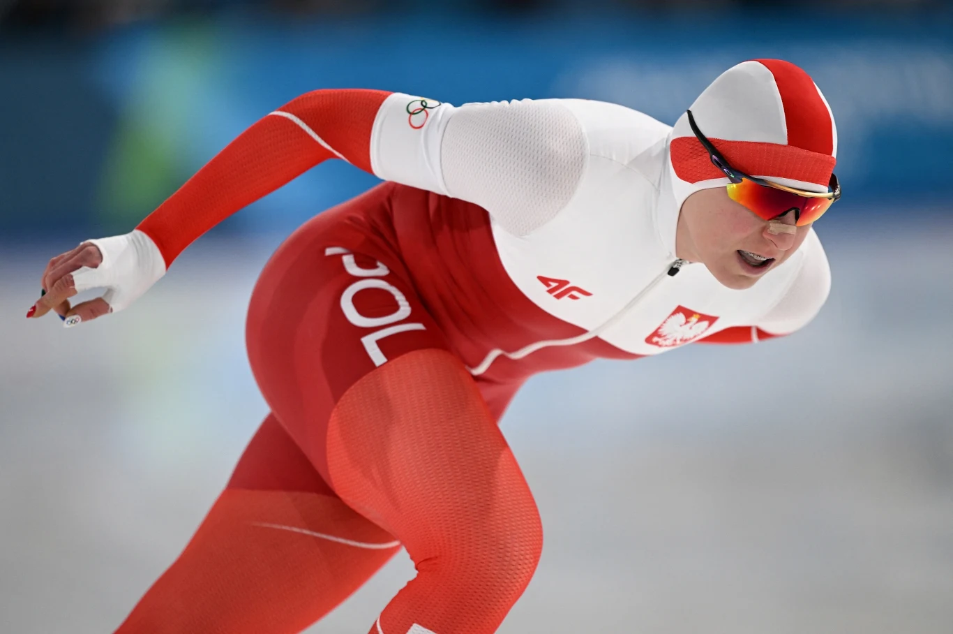 Natalia Czerwonka w akcji Polski panczenista w biało-czerwonym kombinezonie z napisem 'Polska', dynamicznie pochylony podczas wyścigu na lodowisku, w sportowych okularach i kasku w barwach narodowych.