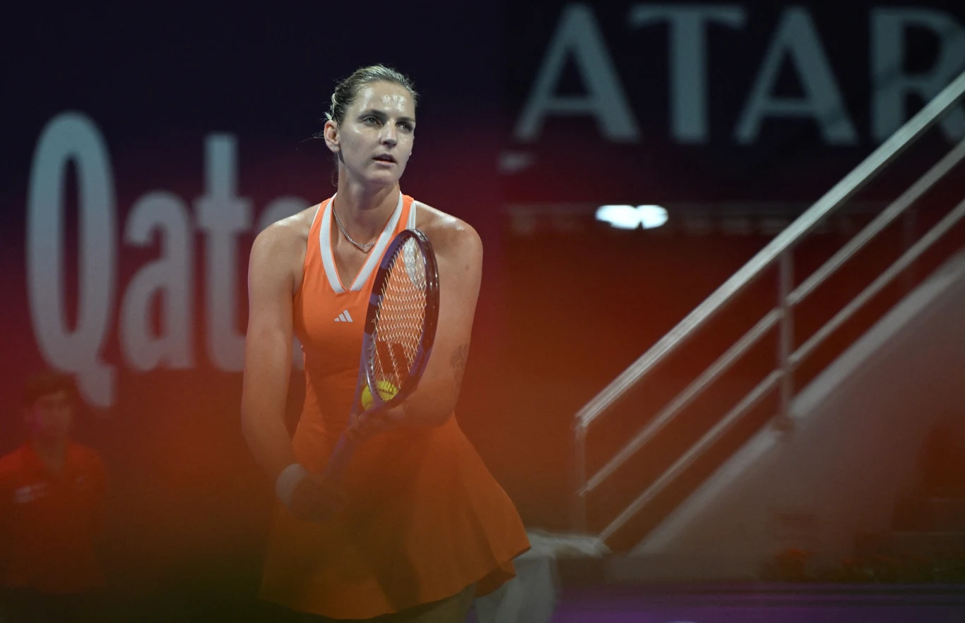 Zawodowa tenisistka w pomarańczowej sukience sportowej stoi na korcie tenisowym z rakietą w ręce, w tle widoczny fragment trybun oraz napisy związane z turniejem w Katarze.