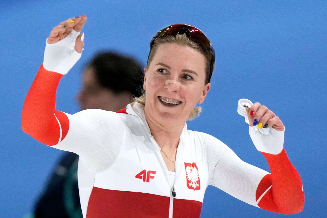 Natalia Czerwonka Sportowiec w biało-czerwonym stroju z godłem Polski na piersi z uniesioną dłonią uśmiecha się, trzymając medal. W tle rozmazana sylwetka innej osoby i niebieskie tło, sugerujące halę sportową.