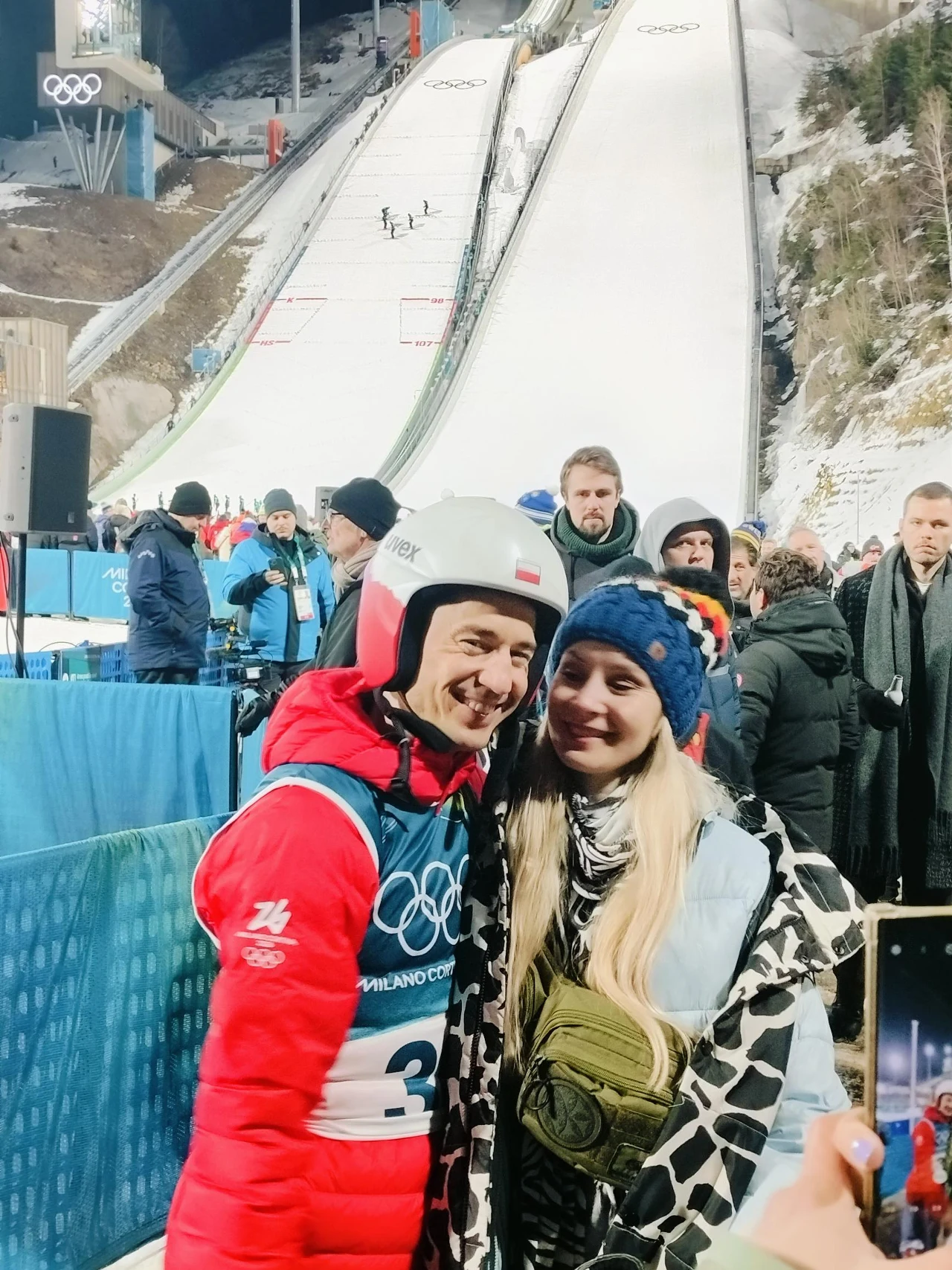 Zadowolony skoczek narciarski ubrany w kombinezon startowy oraz kask pozuje do zdjęcia z uśmiechniętą kobietą na tle skoczni narciarskiej podczas zawodów sportowych. W tle widoczne są tłumy kibiców i sportowa atmosfera zimowych igrzysk.