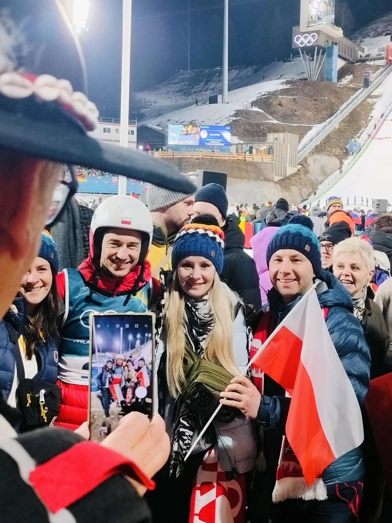 Grupa kibiców wraz ze sportowcem po zawodach zimowych, uśmiechnięci pozują do zdjęcia, jedna z osób trzyma flagę Polski, w tle widać skocznię narciarską oraz tłum innych uczestników wydarzenia.