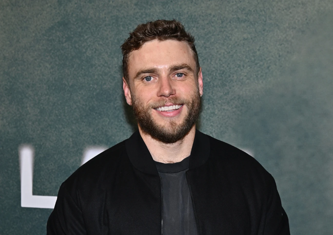 Gus Kenworthy Mężczyzna z jasnymi oczami i brodą ubrany w czarną kurtkę, stoi na tle zielonej ściany z niewyraźnym jasnym napisem w tle, uśmiecha się patrząc w obiektyw.