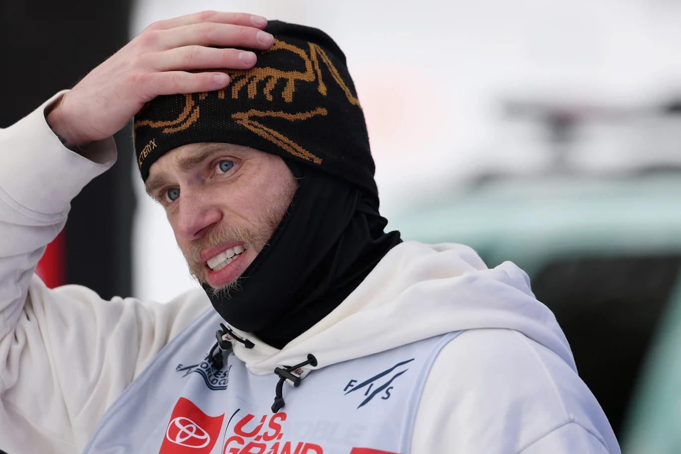 Gus Kenworthy Sportowiec w zimowej czapce i kominiarce, ubrany w biały strój z logotypami sponsorów, trzyma rękę na głowie i wydaje się być zamyślony lub zaniepokojony.