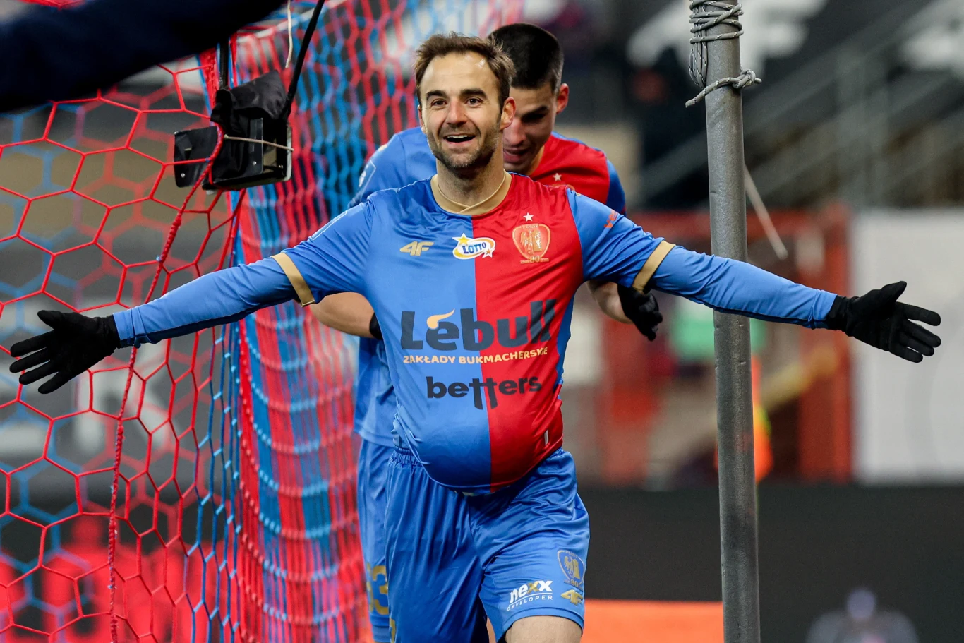 Piłkarz w niebiesko-czerwonym stroju rozkłada ramiona na tle siatki bramki, z widocznym uśmiechem na twarzy, za nim znajduje się inny zawodnik, elementy stadionu w tle, klimat celebracji gola.