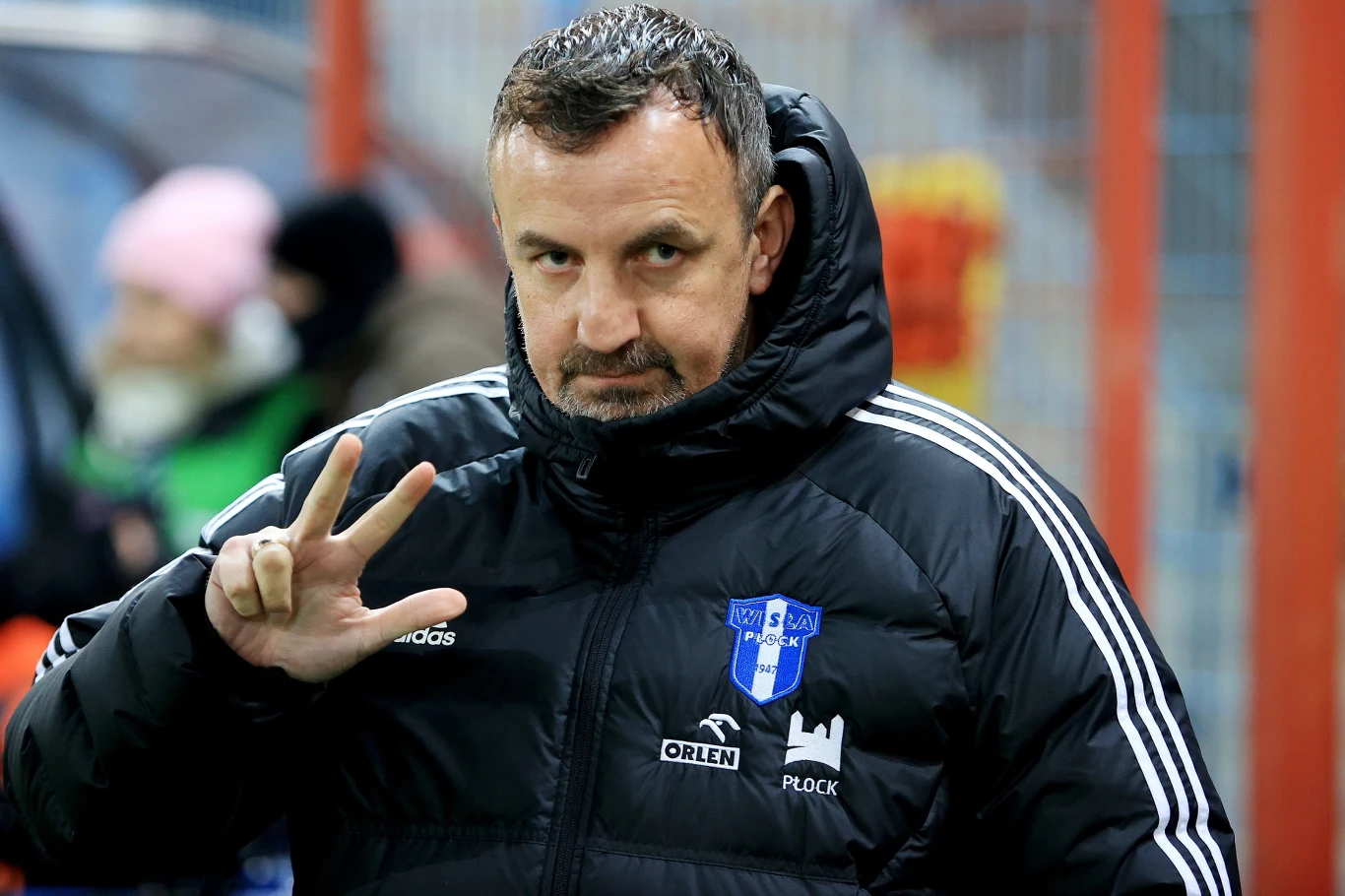 Mężczyzna w kurtce sportowej z herbem klubu Wisła Płock, wykonujący gest trzema palcami, stoi na tle metalowych barierek, w tle widoczne są rozmazane sylwetki innych osób.