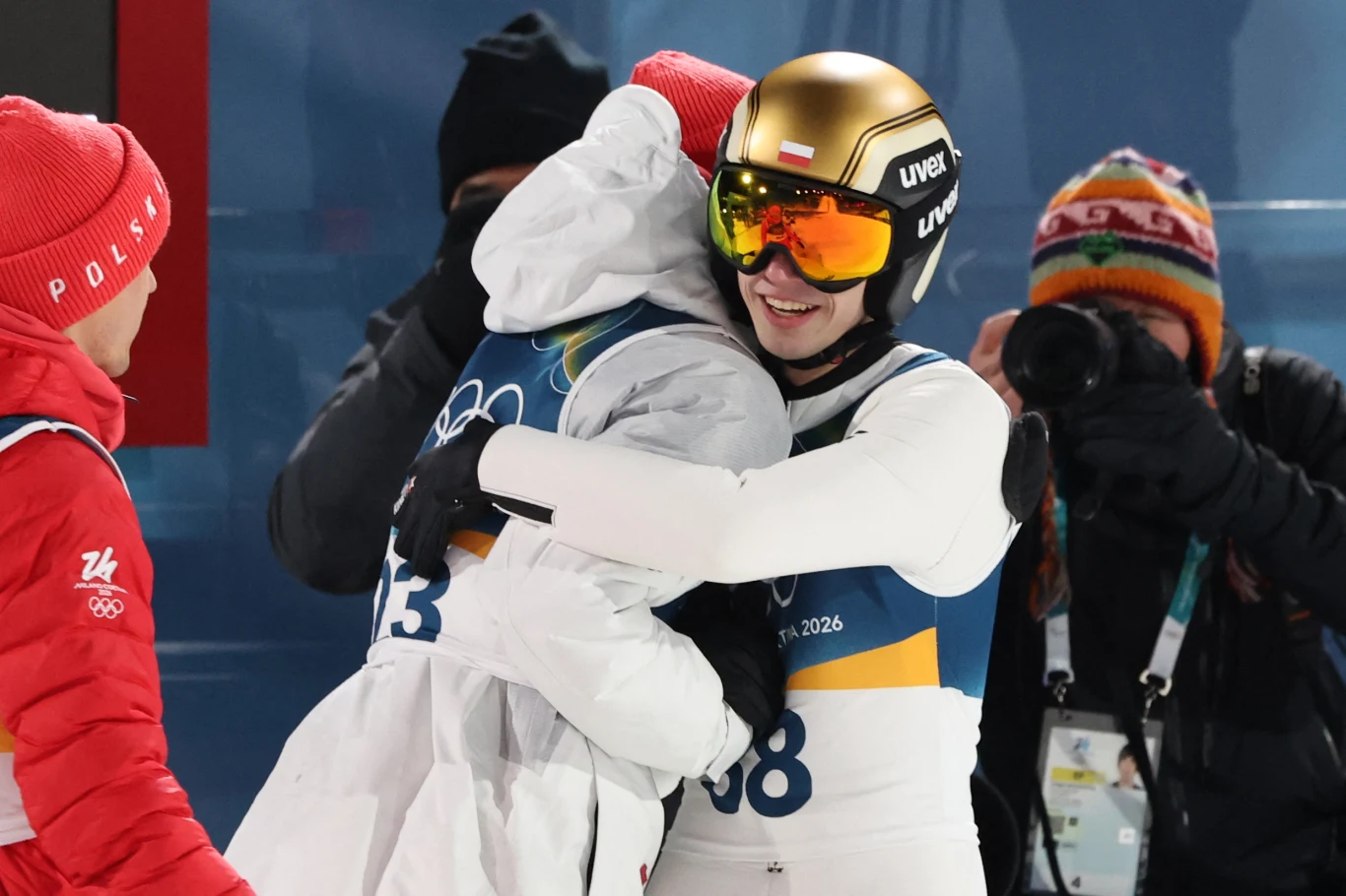 Dwóch sportowców w białych kombinezonach zimowych obejmuje się z radością po zawodach, jeden z nich ma złoty kask i gogle narciarskie z polską flagą, obok znajduje się fotograf w kolorowej czapce oraz inny sportowiec w czerwonej kurtce.