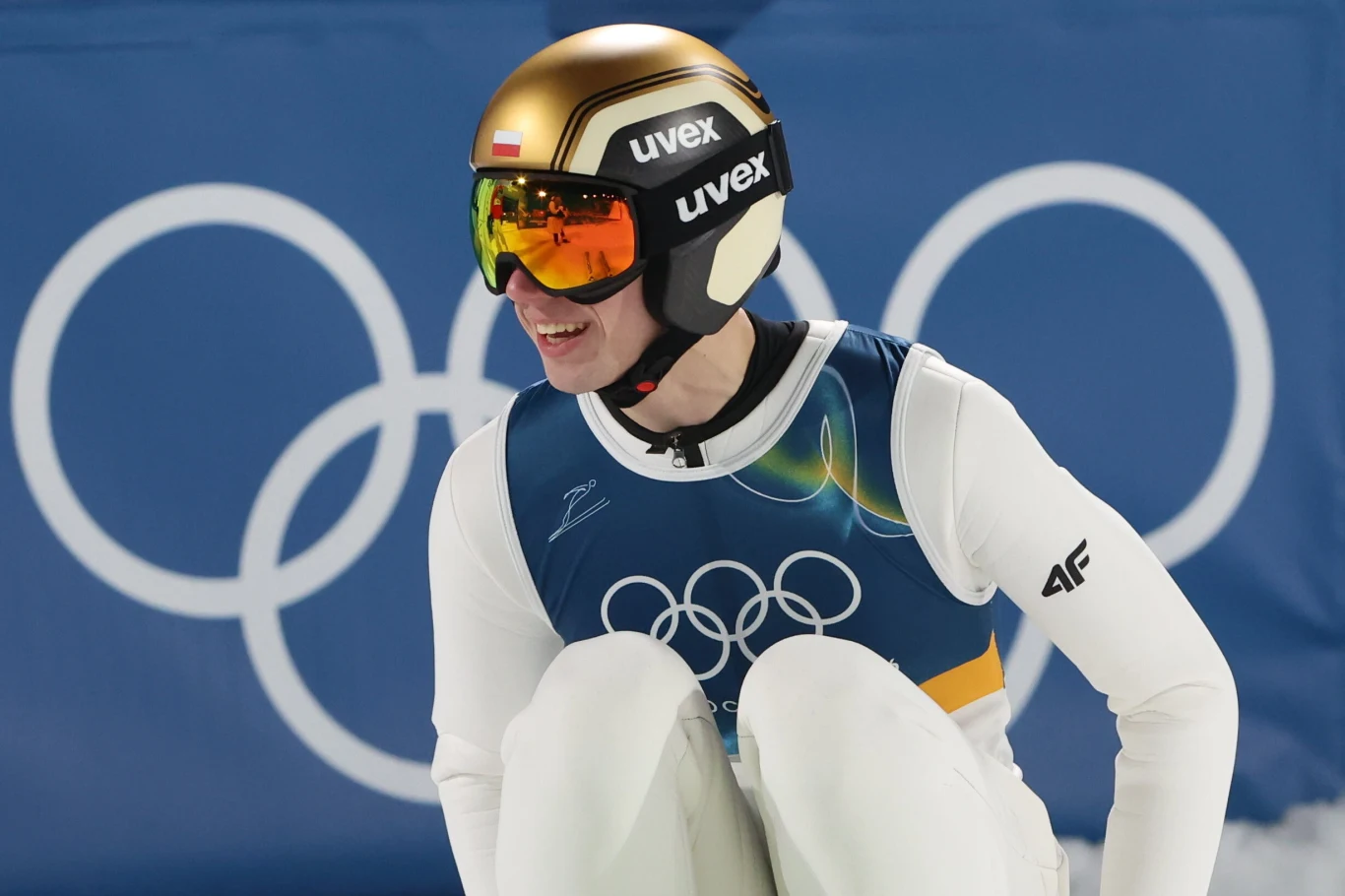 Sportowiec w kombinezonie skoczka narciarskiego, z numerem startowym, kaskiem i goglami, uśmiechnięty, siedzi na tle dużych kół olimpijskich.