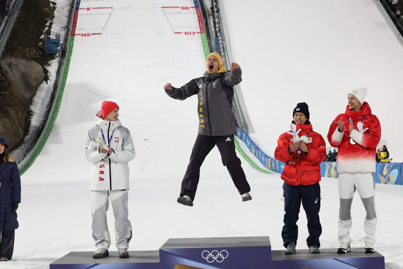 Czterech sportowców stoi na podium podczas ceremonii wręczenia medali na tle skoczni narciarskiej, jedna osoba na najwyższym stopniu wyskakuje w górę z radości, pozostali biją brawo.