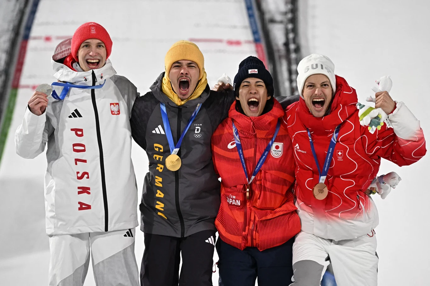 Czterech sportowców w strojach reprezentacyjnych różnych krajów z medalami na szyjach, stojących ramię w ramię na podium, wyrażających radość i entuzjazm po wygranej sportowej.