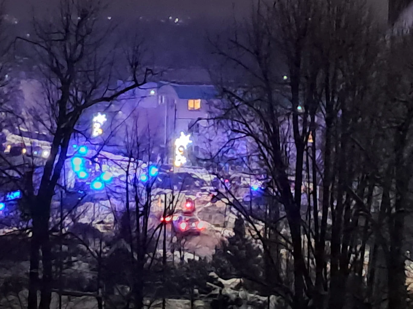 Interwencja policji na "ustawce" pseudokibiców Kilka pojazdów służb ratunkowych z włączonymi sygnałami świetlnymi na ośnieżonej ulicy, otoczonej przez drzewa i udekorowanej świątecznymi iluminacjami, w tle widoczny budynek mieszkalny z zapalonym światłem.