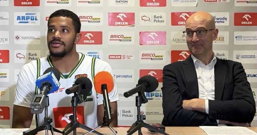 Dwóch mężczyzn siedzi przy stole konferencyjnym, przed nimi ustawione są mikrofony. Po lewej sportowiec w koszulce drużyny koszykarskiej, po prawej mężczyzna w garniturze i okularach z założonymi rękami. W tle widoczna jest ściana z logotypami sponsorów.