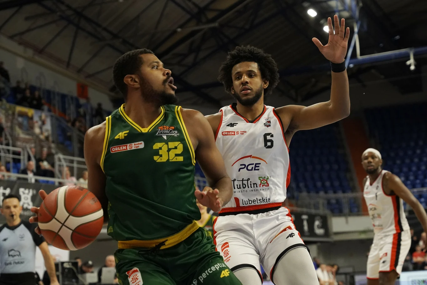 Koszykarz w zielonym stroju z numerem 32 drybluje piłką podczas meczu, próbując przedrzeć się przez obronę zawodnika w białym stroju z numerem 6. W tle widoczna hala sportowa z publicznością.