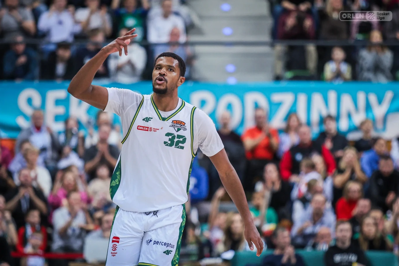 Koszykarz w białym stroju z numerem 32 wykonuje gest ręką podczas meczu, w tle widać tłum kibiców na trybunach.