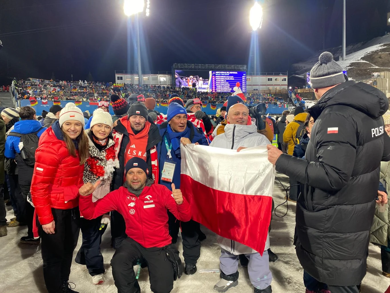 Grupa kibiców ubrana w zimowe stroje i w barwy narodowe Polski świętuje na stadionie sportowym, trzymając dużą flagę Polski. W tle tłum widzów na trybunach oraz jasne oświetlenie stadionu.