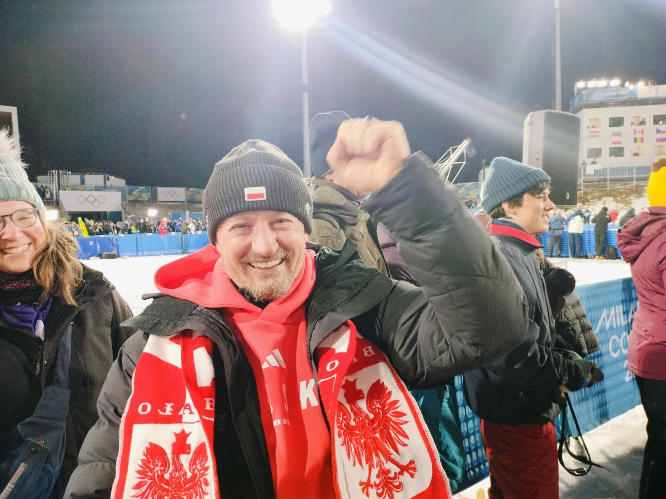 Uśmiechnięty mężczyzna ubrany w zimową kurtkę oraz szalik z wizerunkiem białego orła i napisem 'POLSKA', stojący na tle trybun podczas zimowej imprezy sportowej, unoszący zaciśniętą pięść w geście radości.
