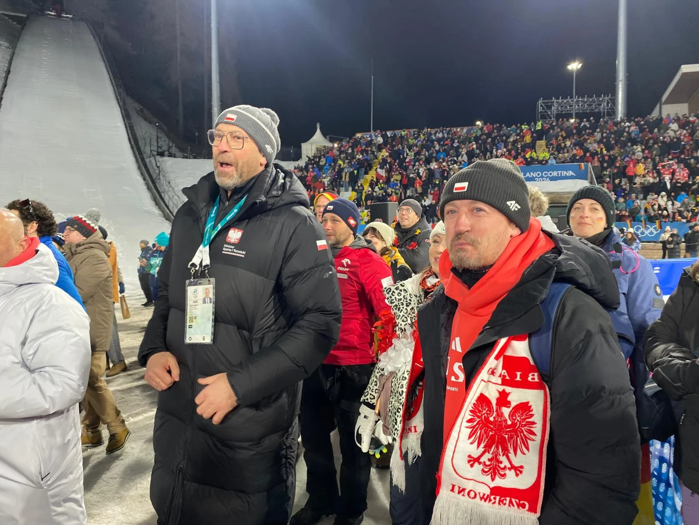 Grupa kibiców ubranych w zimowe kurtki oraz czerwono-białe szaliki z orłem i napisem Polska, stojących na tle skoczni narciarskiej oraz trybun wypełnionych publicznością podczas zawodów sportowych nocą.