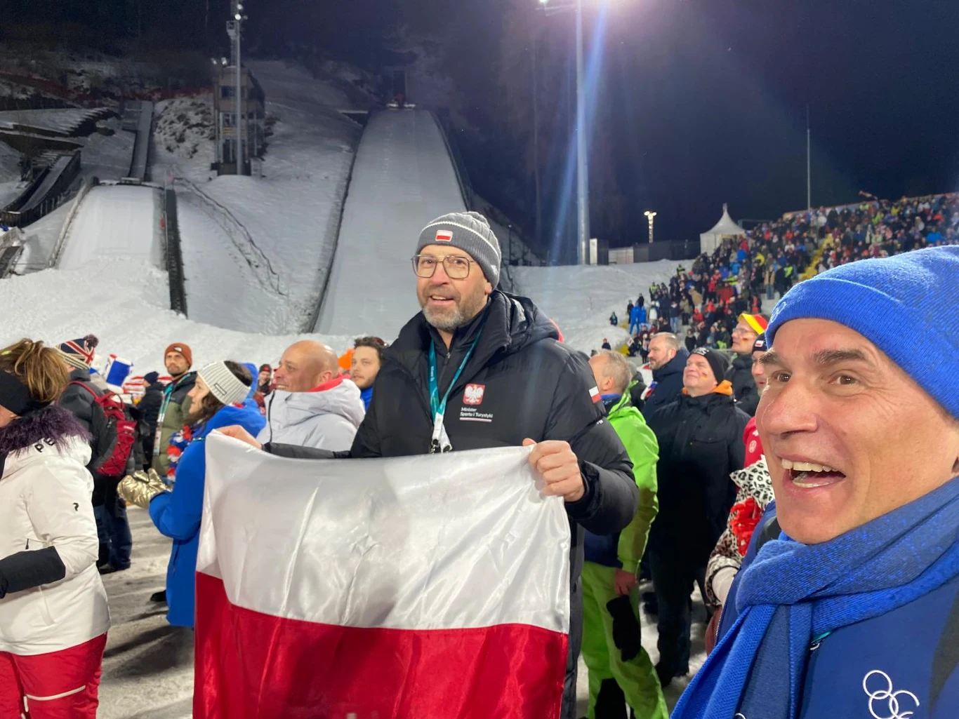 Tłum ludzi na skoczni narciarskiej, mężczyzna na pierwszym planie trzyma polską flagę, w tle kibice oraz oświetlona skocznia, zimowa atmosfera, wszyscy ubrani w ciepłe kurtki.