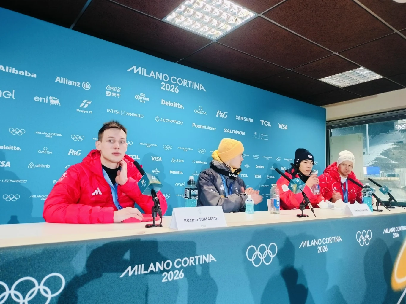 Grupa sportowców w czerwonych kurtkach siedzących przy stole konferencyjnym na tle ściany z logotypami sponsorów oraz napisem Milano Cortina 2026, po lewej stronie mikrofony i tabliczki z nazwiskami.