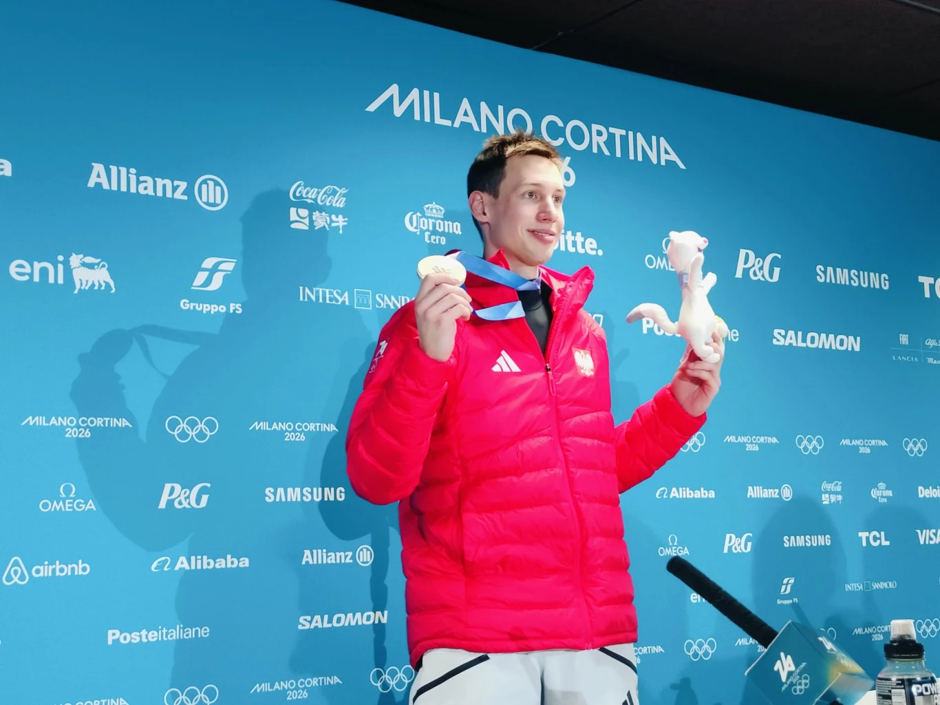 Kacper Tomasiak Sportowiec w czerwonej kurtce trzyma medal olimpijski i maskotkę, stojąc na tle ścianki z logotypami Milano Cortina i sponsorów. Uśmiechnięty mężczyzna pozuje do zdjęcia po zdobyciu nagrody.