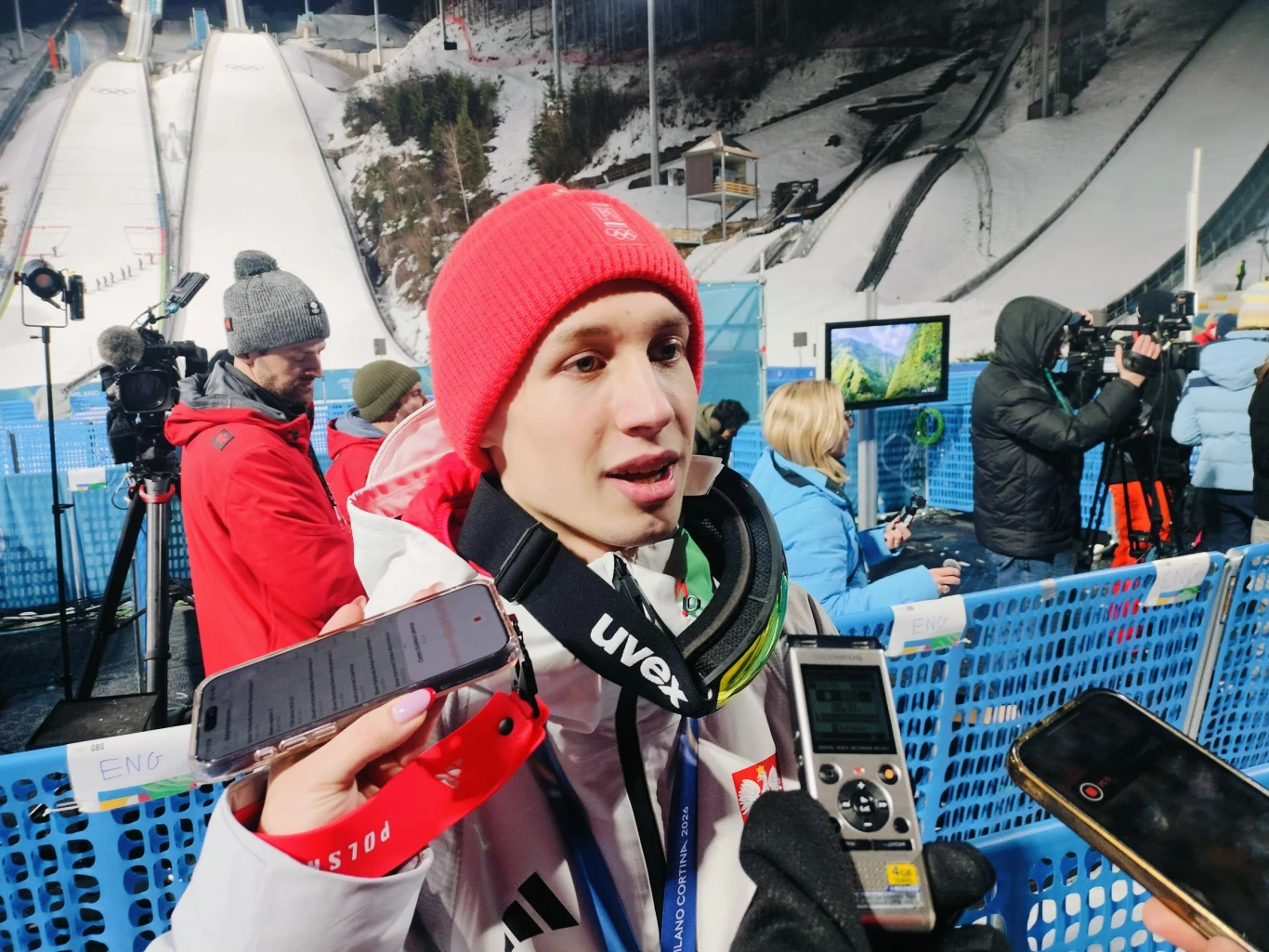 Kacper Tomasiak Młody skoczek narciarski w czerwonej czapce i białej kurtce udziela wywiadu dziennikarzom po zawodach, wokół liczne mikrofony i dyktafony, w tle ośnieżona skocznia oraz ekipy telewizyjne.