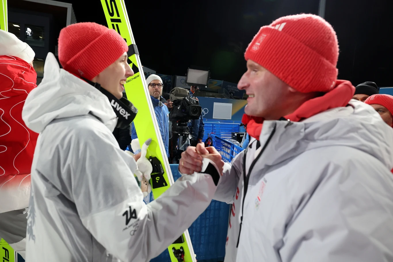 Sportowcy ubrani w białe kurtki i czerwone czapki gratulują sobie po zawodach narciarskich; w tle widać duże narty i kamerzystę rejestrującego wydarzenie.