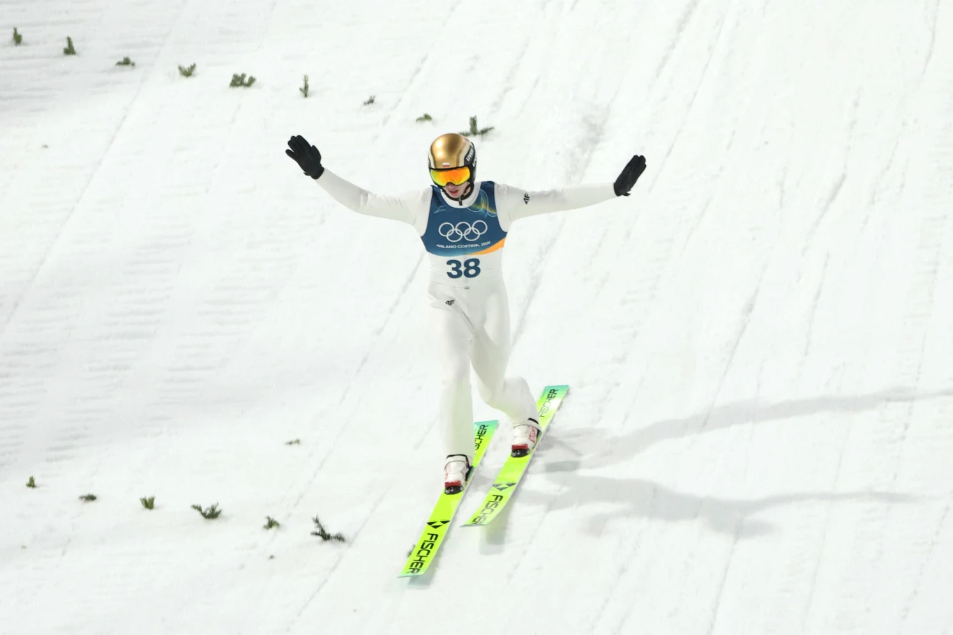 Skoczek narciarski w trakcie lądowania na skoczni, ubrany w biały kombinezon i złoty kask, unosi ręce w geście triumfu. W tle widoczne ślady nart na śniegu oraz fragmenty zielonej dekoracji.