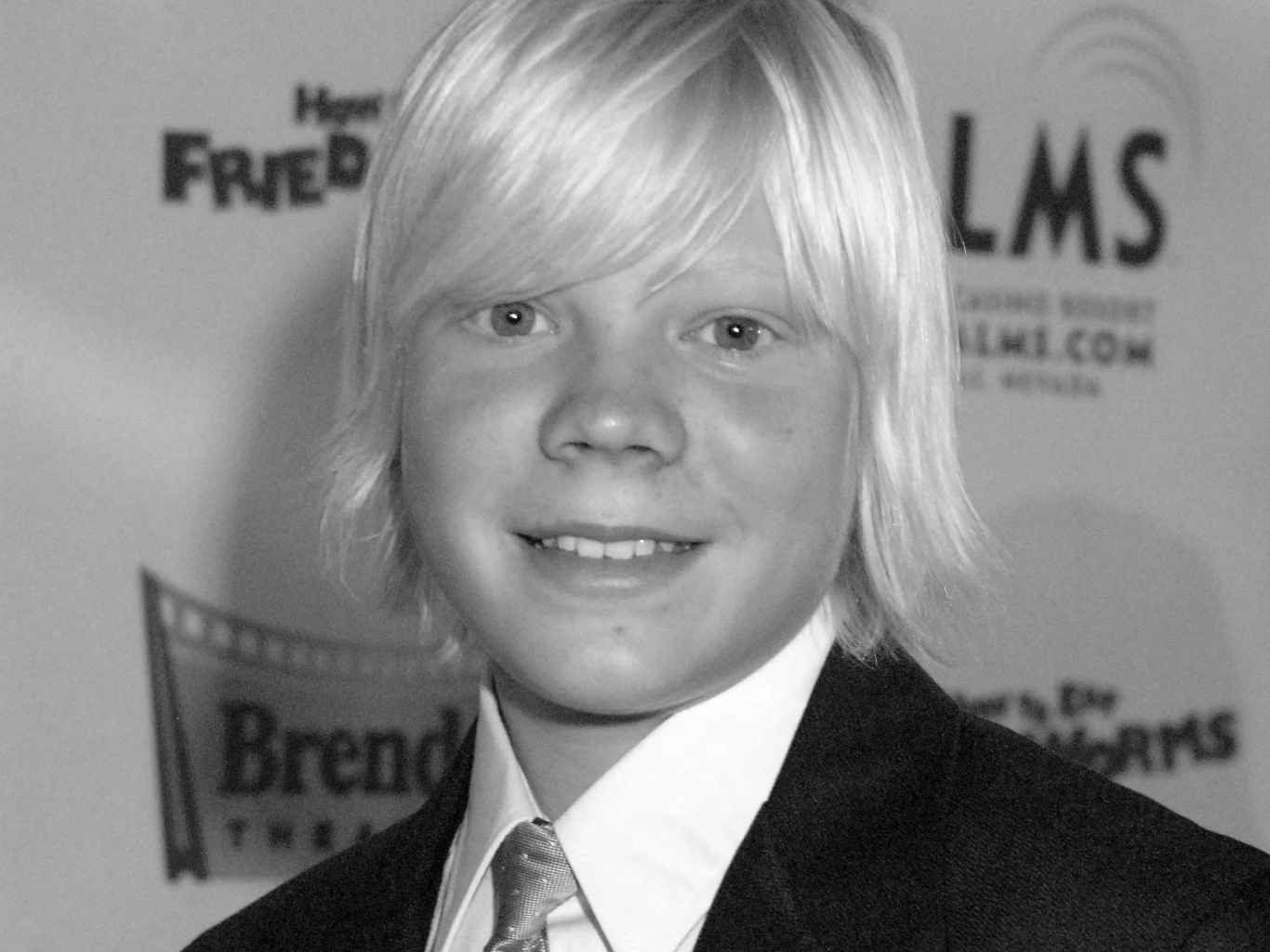 Blake Garrett w 2006 roku Młody chłopak z jasnymi włosami uśmiecha się, ubrany elegancko w garnitur i krawat, w tle rozmazane napisy i banery sugerujące udział w wydarzeniu lub premierze filmowej.