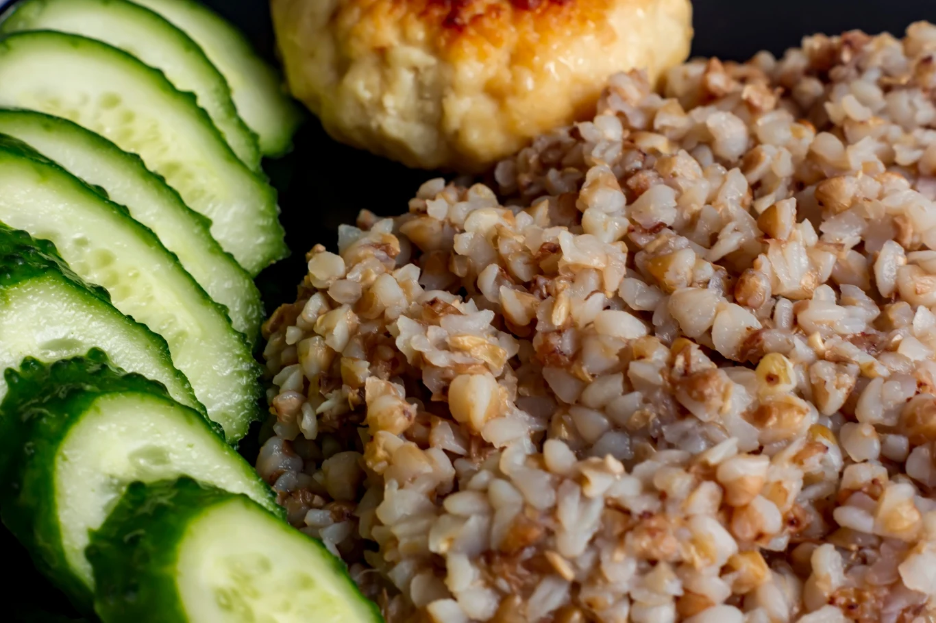 Plasterki świeżego ogórka ułożone obok porcji ugotowanej kaszy gryczanej i zapiekanej kotleciki z mięsa drobiowego.