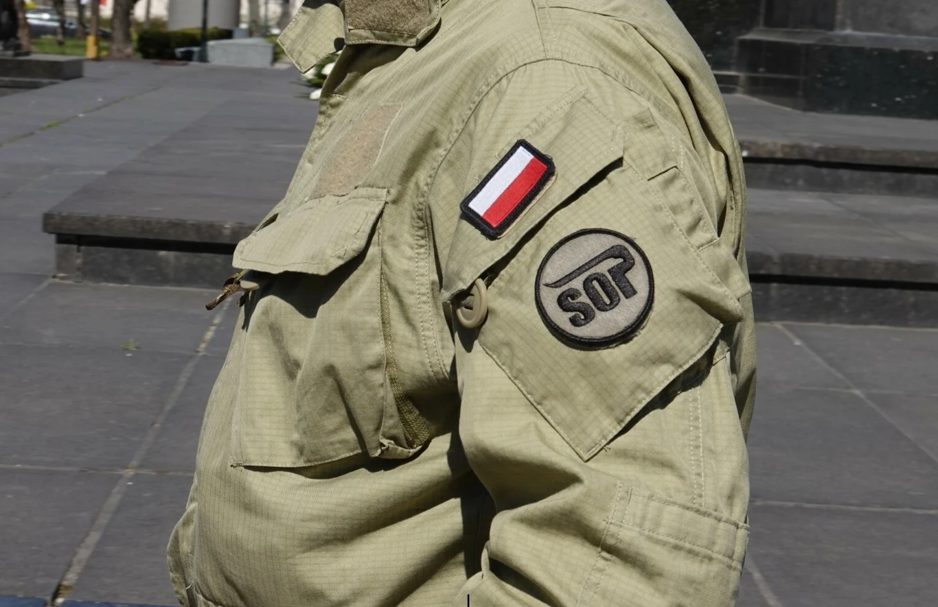 Fragment sylwetki osoby w mundurze z naszywkami SOP oraz flagą Polski, wykonany w scenerii miejskiej, bez widocznej twarzy.