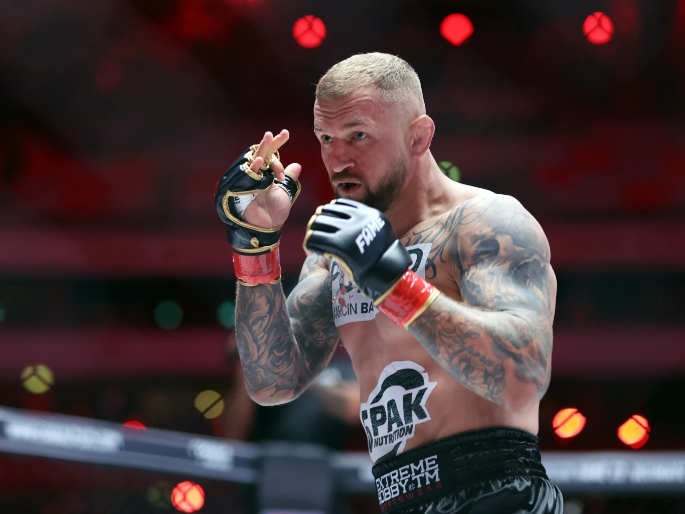 Marcin Wrzosek Zawodnik MMA w ringu, ubrany w sportowe szorty i rękawice, z tatuażami na ramionach, skoncentrowany i przyjmujący pozycję bojową, w tle rozświetlona hala.