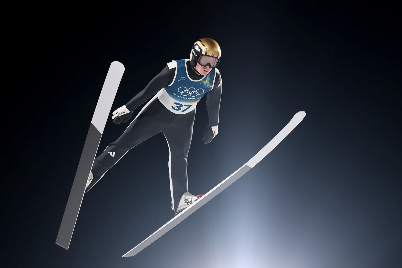 Skoczek narciarski w powietrzu w trakcie lotu, ubrany w czarny kombinezon z numerem startowym 37 i kaskiem, na tle ciemnego nieba i podświetlenia, widoczne logo igrzysk olimpijskich na kamizelce.