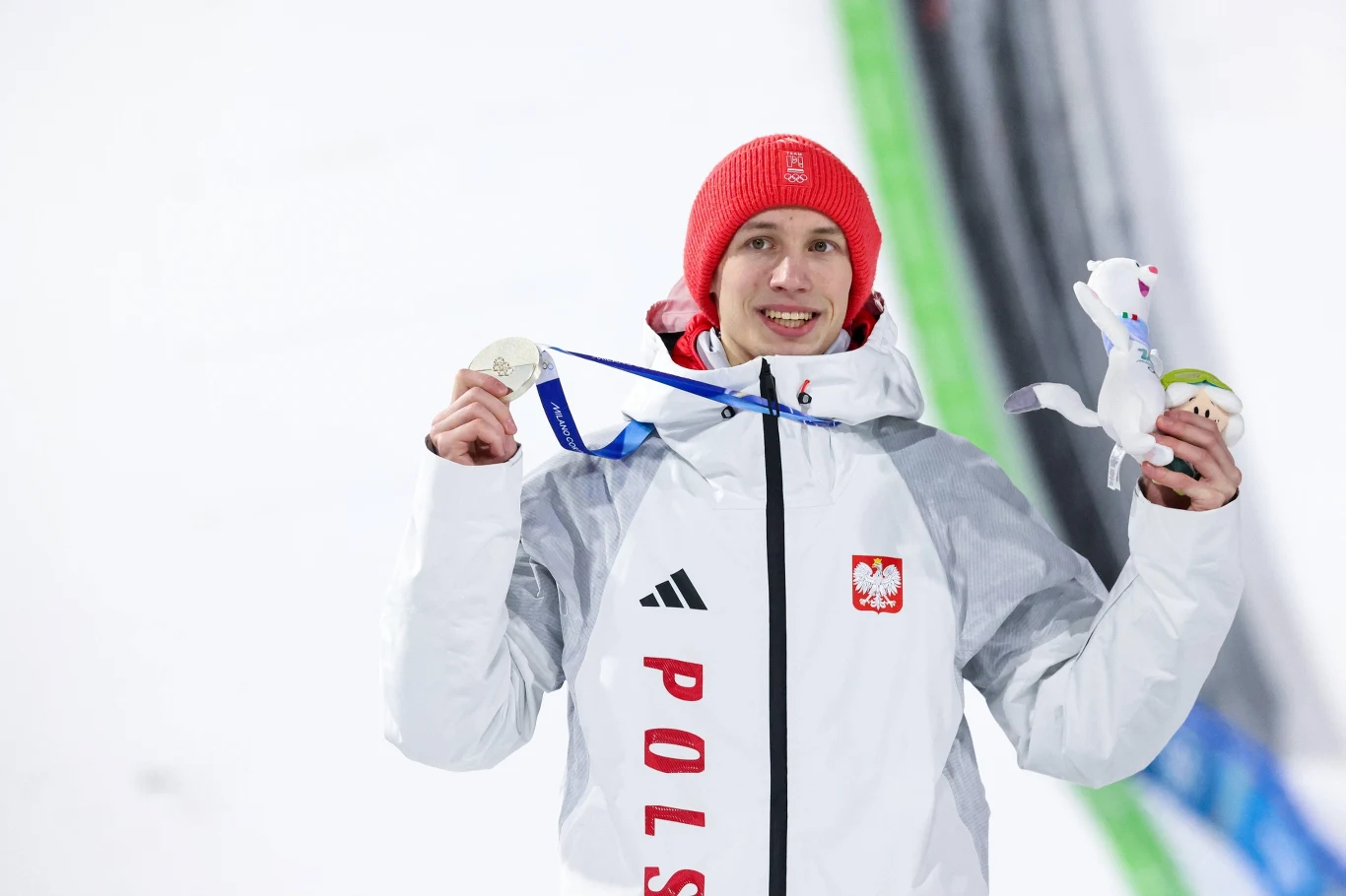 Kacper Tomasiak Sportowiec w białej kurtce z napisem 'Polska' i czerwonej czapce trzyma srebrny medal oraz maskotkę, uśmiechając się do kamery, w tle widoczny fragment skoczni zimowej.