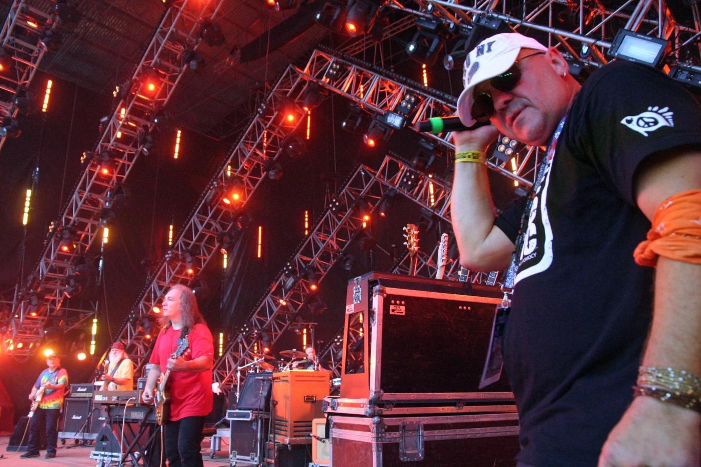 Zespół muzyczny występujący na dużej scenie, kolorowe światła sceniczne, widoczny wokalista z mikrofonem na pierwszym planie, muzycy grający na gitarach i instrumentach klawiszowych w tle, profesjonalny sprzęt nagłośnieniowy i konstrukcja sceniczna.