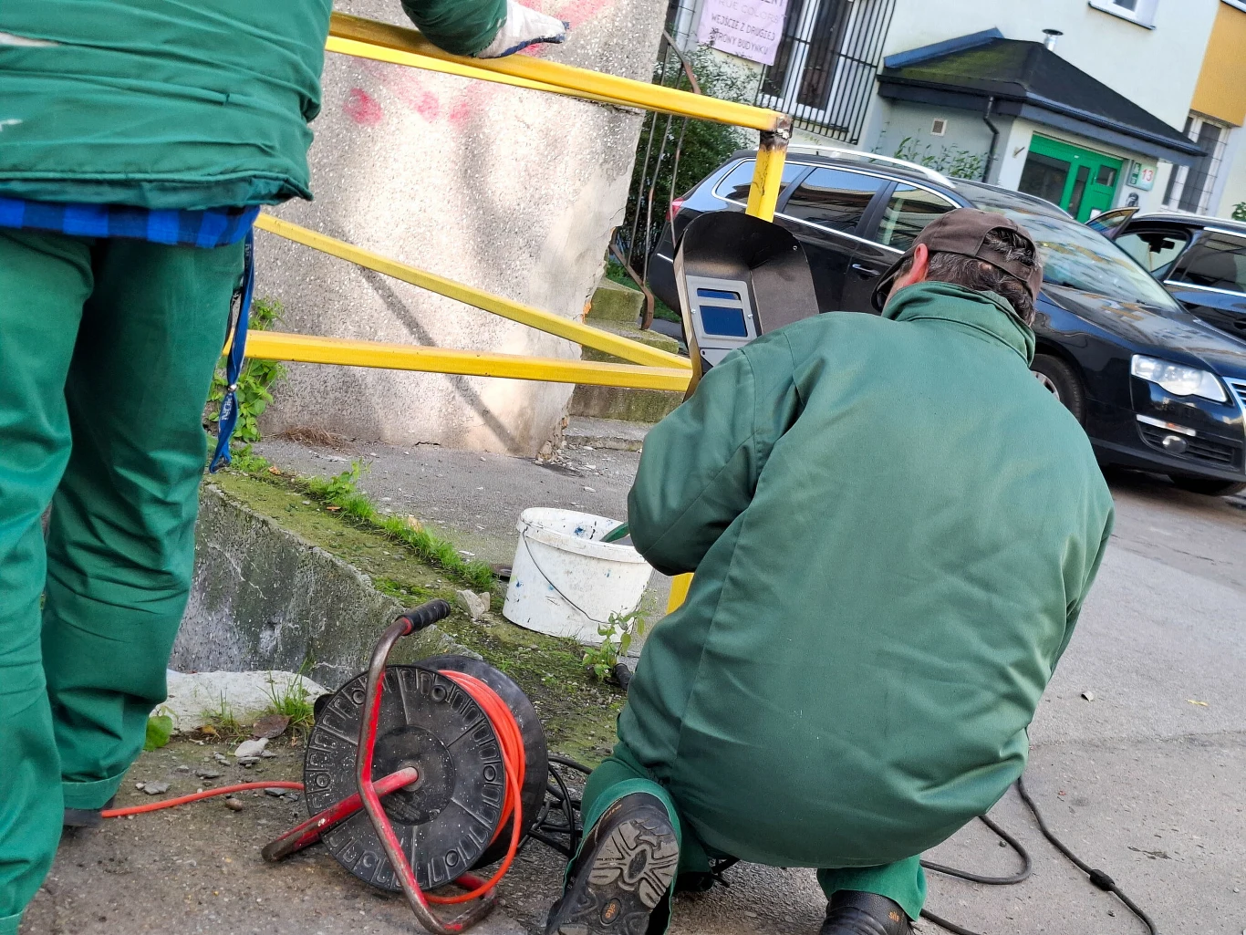 W ostatnim dniu sierpnia 2025 r. pracę w Polsce wykonywało 1103,1 tys. cudzoziemców - podał GUS Dwóch pracowników ubranych w zielone kombinezony wykonuje prace techniczne przy żółtych barierkach na chodniku, obok znajduje się zwinięty kabel elektryczny i biały pojemnik.