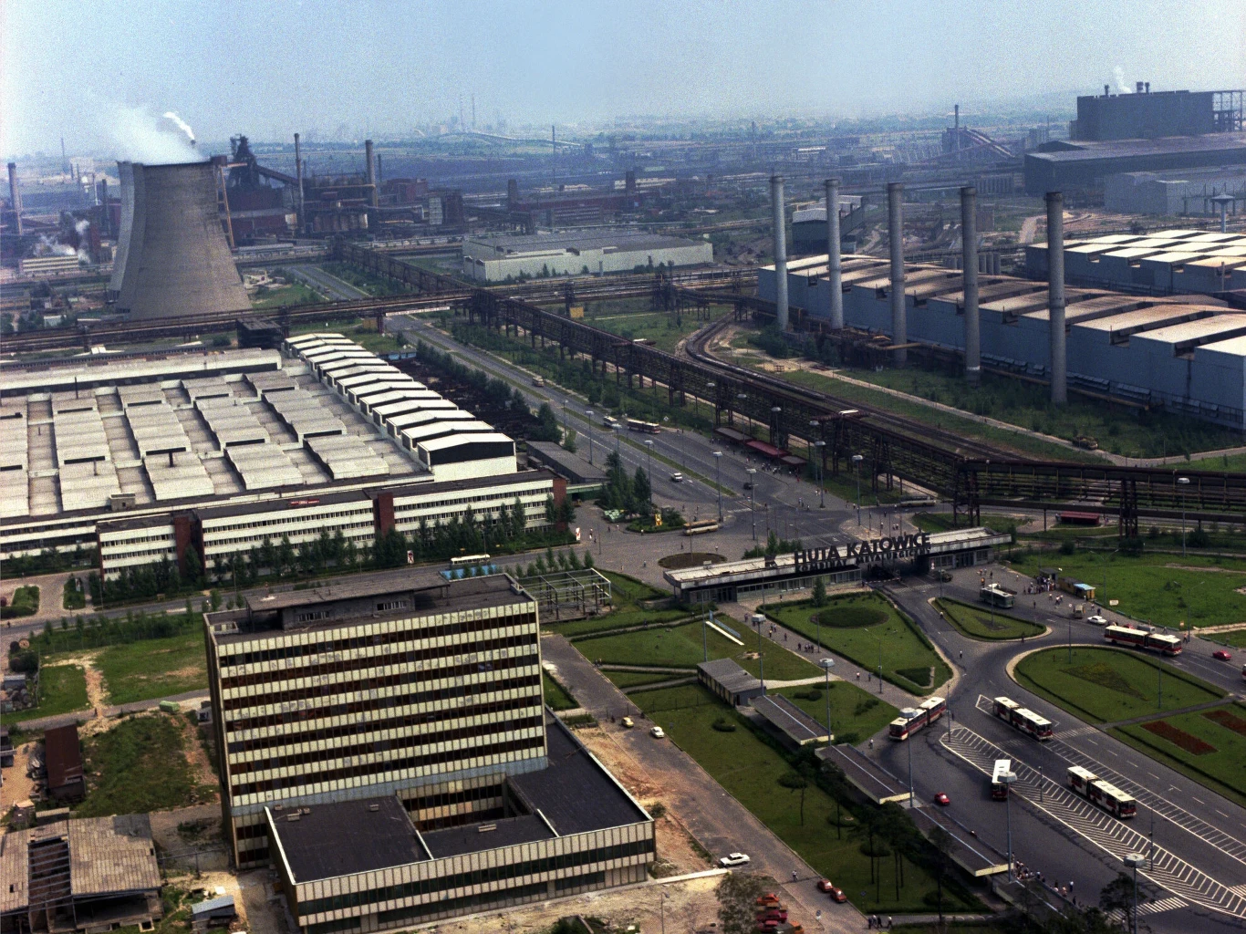 Widok na Hutę Katowice z lotu ptaka, ok. 1986 Kompleks przemysłowy z dużą liczbą hal i kominów, biurowiec na pierwszym planie, szerokie drogi i skrzyżowania, w tle rozległy krajobraz miejski z licznymi instalacjami przemysłowymi oraz chłodnią kominową.