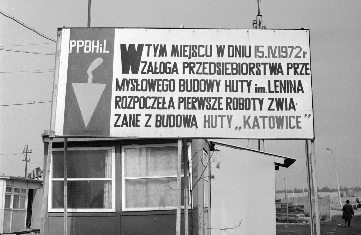 Tablica upamiętniająca budowę Huty Katowice, 1974 rok Tablica informacyjna upamiętniająca rozpoczęcie prac związanych z budową Huty Katowice przez przedsiębiorstwo budowy huty imienia Lenina w dniu 15 kwietnia 1972 roku, umieszczona na placu budowy, w tle fragment zaplecza budowy i pusty teren.