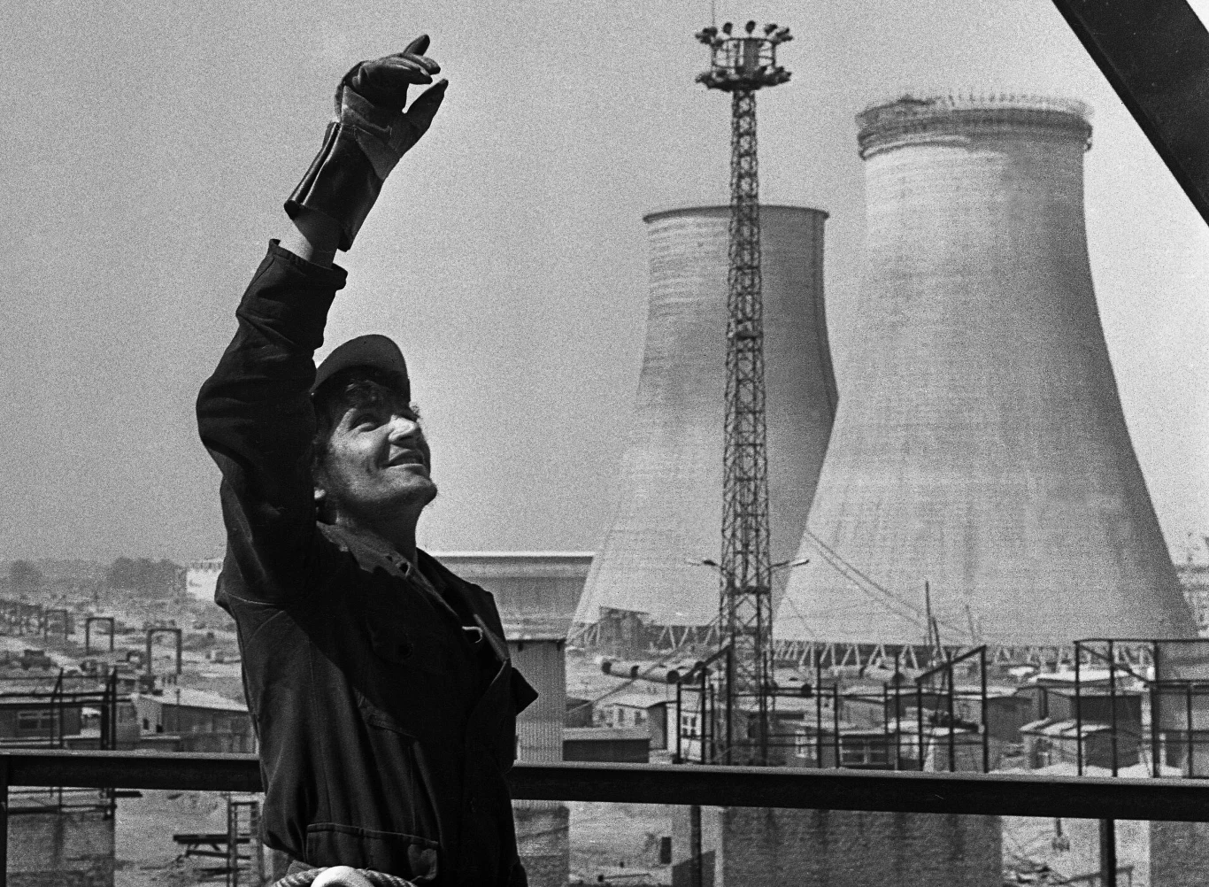 Budowa Huty Katowice, ok. 1974 rok Pracownik przemysłowy w kombinezonie i rękawicach unosi rękę w górę, w tle widoczne potężne chłodnie kominowe oraz infrastruktura zakładu przemysłowego. Sceneria miejska skupiona na obszarze fabrycznym.