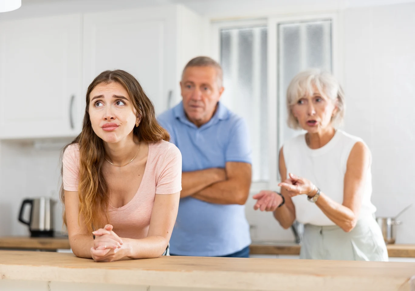 Młoda kobieta z wyrazem frustracji lub zniecierpliwienia na twarzy stoi przy kuchennym blacie, za nią stoi starsze małżeństwo, które wydaje się prowadzić z nią żywą dyskusję lub kłótnię