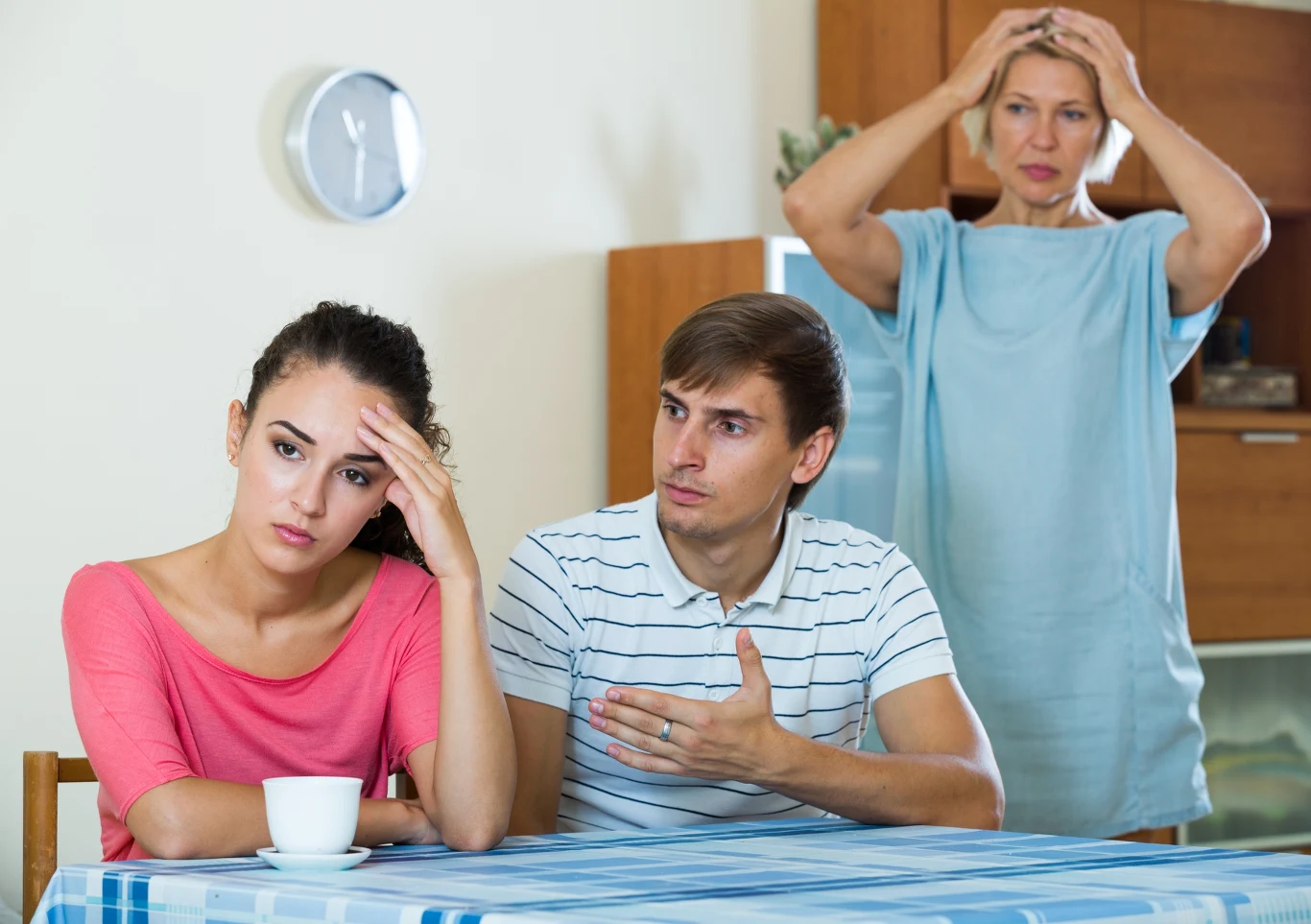 Kobieta siedzi przy stole z wyrazem zmartwienia na twarzy, obok niej mężczyzna gestykuluje, próbując coś wyjaśnić, w tle starsza kobieta trzyma się za głowę ze zmartwieniem.