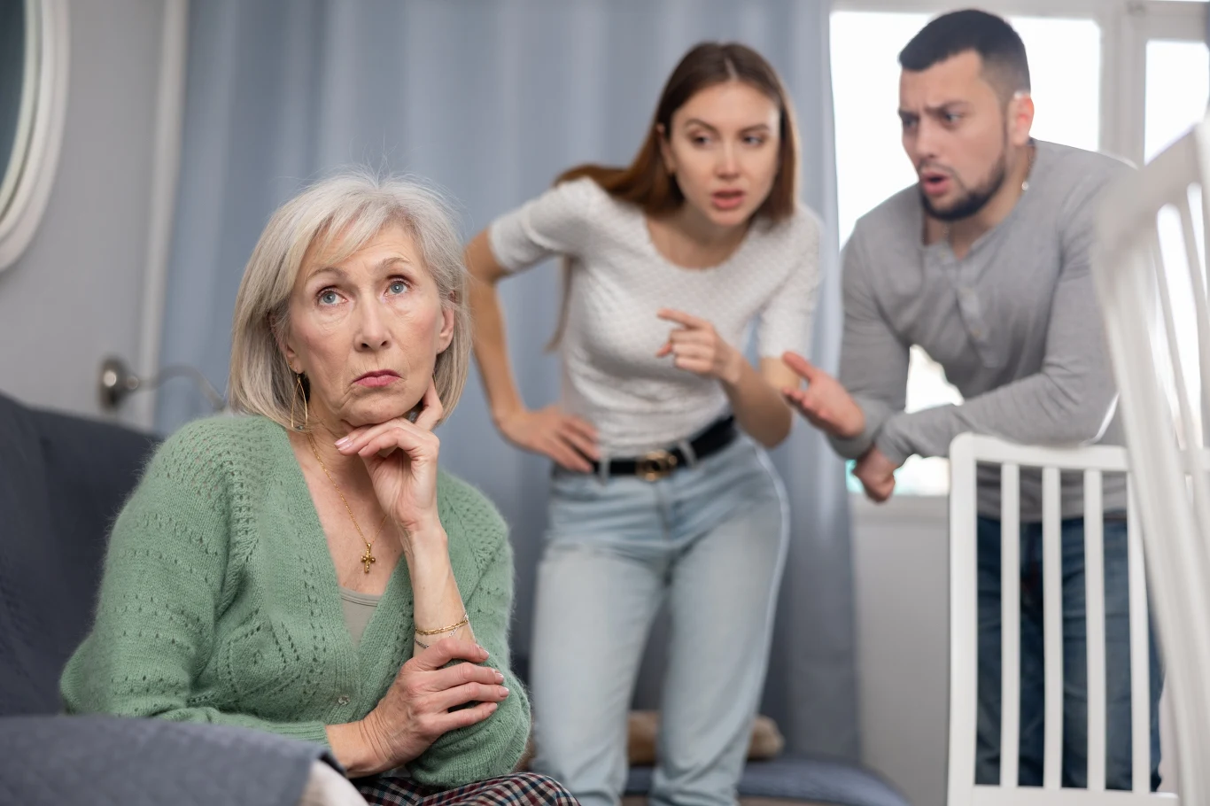Starsza kobieta siedzi na kanapie z zamyślonym, znudzonym wyrazem twarzy, podczas gdy młoda para w tle gestykuluje i wyraża silne emocje, prawdopodobnie podczas kłótni lub nieporozumienia.