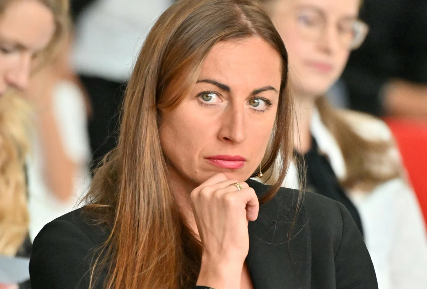 Kobieta o długich brązowych włosach w czarnym stroju, podpierająca brodę dłonią i patrząca z powagą przed siebie, w tle rozmyci ludzie.