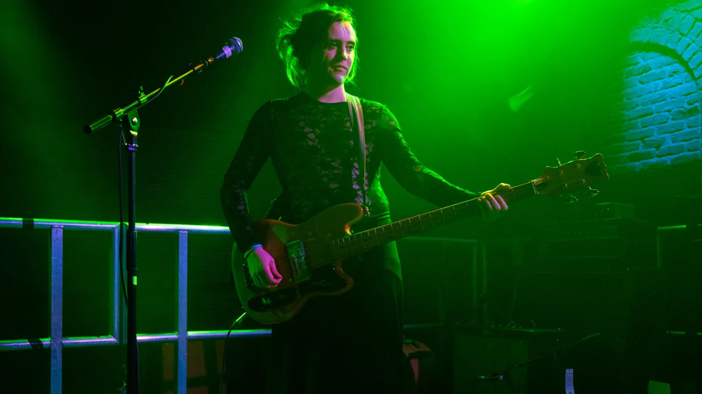 Jamie Hyland (M(h)aol) Muzyk grający na gitarze basowej na scenie, oświetlony zielonym światłem, z widocznym mikrofonem i fragmentem ceglanej ściany w tle.