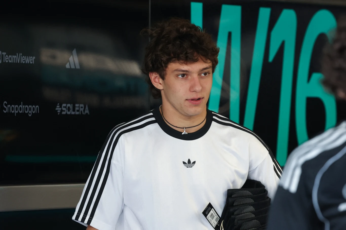 Młody mężczyzna w białej koszulce sportowej z logiem Adidas, trzymający czarną odzież, stoi na tle ciemnej tablicy z dużymi literami i logotypami firm.