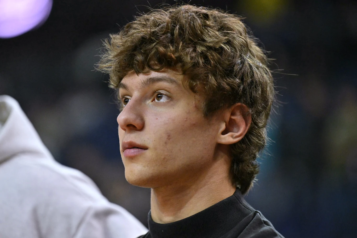 Młody mężczyzna z kręconymi brązowymi włosami patrzący lekko w górę, ubrany w czarną bluzę, neutralne tło sprawia, że cała uwaga skupia się na jego twarzy i wyrazie zamyślenia.