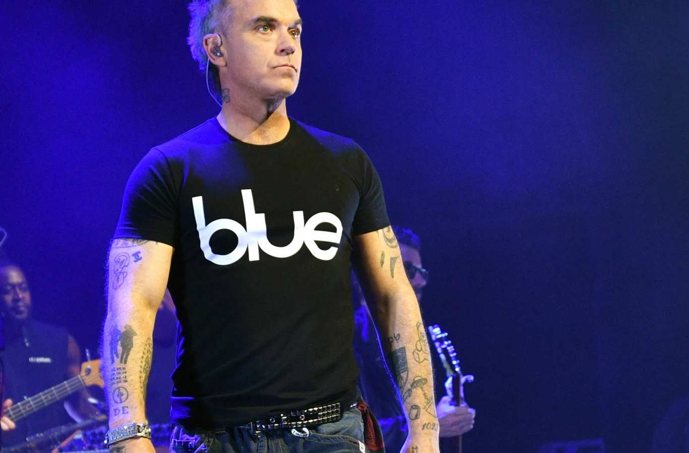 Robbie Williams Mężczyzna z tatuażami na przedramionach ubrany w czarną koszulkę z napisem „blue” stoi na scenie podczas koncertu, w tle widoczni muzycy i niebieskie światła.