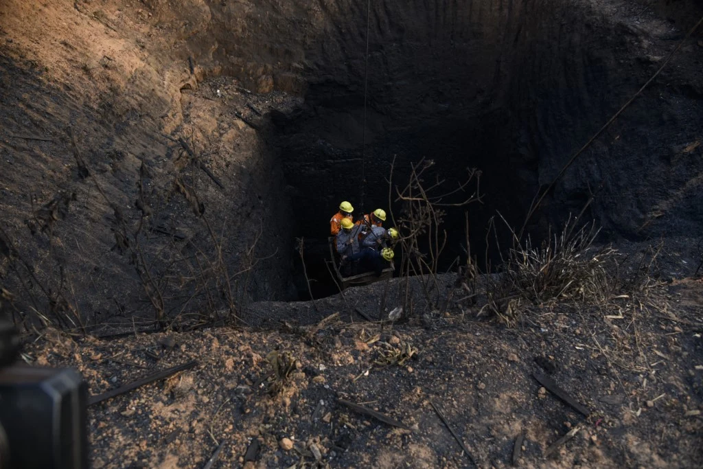 Dziesiątki ofiar wybuchu w nielegalnej kopalni w Indiach. Pracowali w skandalicznych warunkach Trzech ratowników w kaskach ochronnych schodzących do głębokiego, ciemnego szybu na wypalonej, spalonej powierzchni terenu. Wokół leży wypalona ziemia oraz zeschłe gałęzie.
