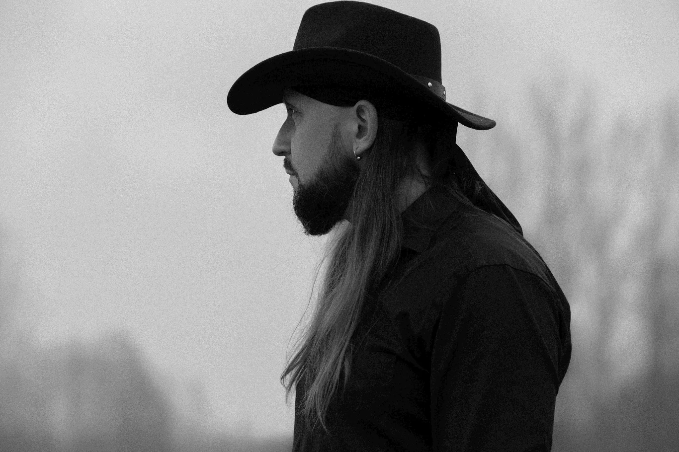 Ukć to solowy projekt Łukasza "Icanraza" Sarnackiego Mężczyzna z długimi włosami i brodą ubrany w czarną koszulę oraz kapelusz, patrzący w bok na tle rozmytego, szarego krajobrazu.